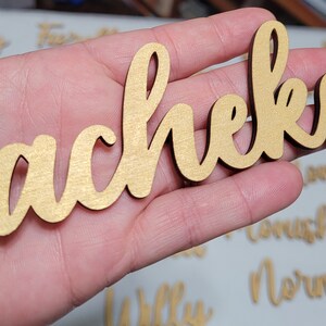 May include: A gold-colored wooden cutout featuring the name Sacheka in a cursive script font. The letters are connected in a single piece with a smooth, metallic-toned finish on the front surface and dark edges, showcasing a decorative name sign.