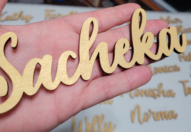 May include: A gold-colored wooden cutout featuring the name Sacheka in a cursive script font. The letters are connected in a single piece with a smooth, metallic-toned finish on the front surface and dark edges, showcasing a decorative name sign.