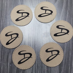 May include: Six round beige coasters with a black stylised letter "D" design. The coasters are arranged in a circle on a wooden surface.