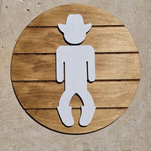 May include: A circular wooden sign featuring a dark brown stained finish with horizontal grooves that mimic planking. Centered on the sign is a white, flat, cutout silhouette of a figure wearing a cowboy hat and boots, standing in a bow-legged stance.