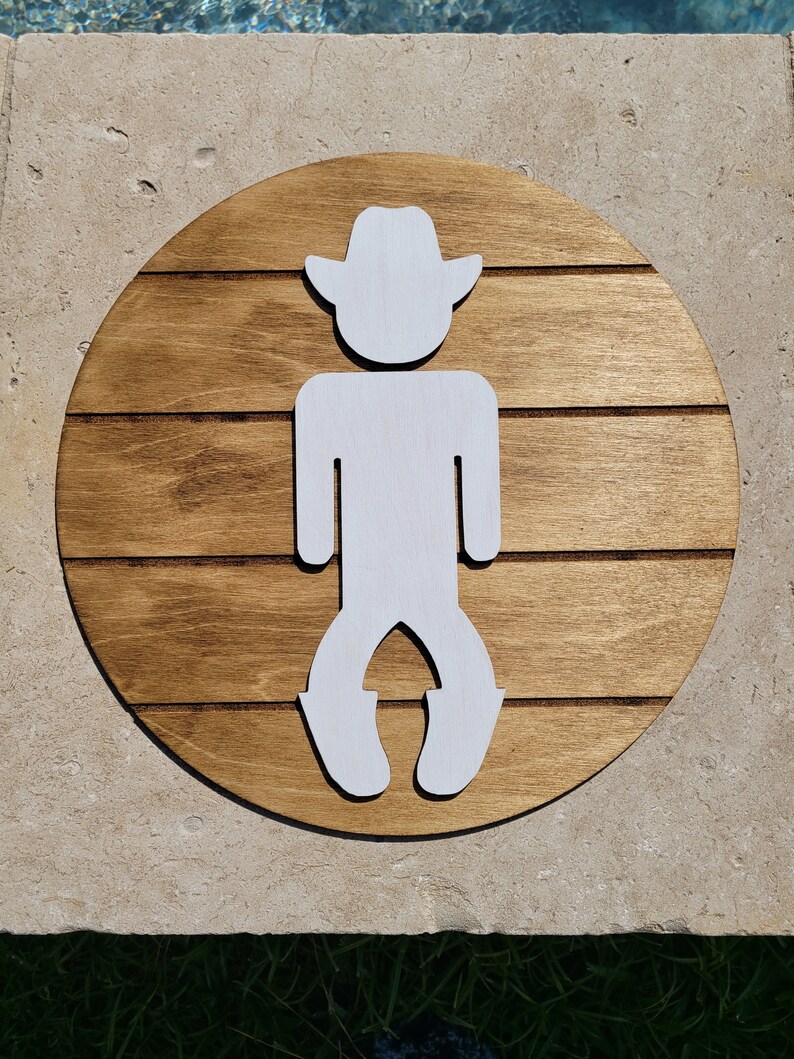 May include: A circular wooden sign featuring a dark brown stained finish with horizontal grooves that mimic planking. Centered on the sign is a white, flat, cutout silhouette of a figure wearing a cowboy hat and boots, standing in a bow-legged stance.