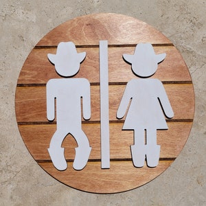 May include: A circular wooden sign featuring horizontal slats in a medium brown finish. Attached to the front are two white, flat, cutout figures wearing cowboy hats and boots, separated by a vertical white bar, representing restroom signage.