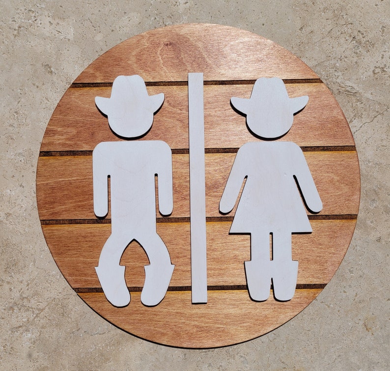 May include: A circular wooden sign featuring horizontal slats in a medium brown finish. Attached to the front are two white, flat, cutout figures wearing cowboy hats and boots, separated by a vertical white bar, representing restroom signage.