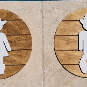 May include: Two circular wooden signs with a dark brown stained finish and horizontal plank detailing. Each sign features a white cutout figure wearing a cowboy hat. One figure wears a skirt and boots, while the other wears pants and boots.