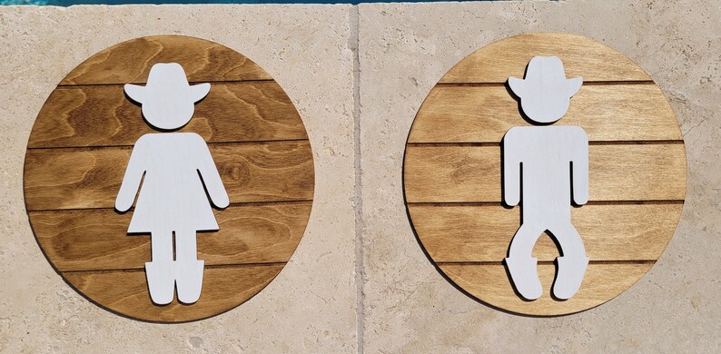 May include: Two circular wooden signs with a dark brown stained finish and horizontal plank detailing. Each sign features a white cutout figure wearing a cowboy hat. One figure wears a skirt and boots, while the other wears pants and boots.