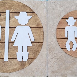 May include: Two circular wooden signs featuring white cutout figures of a cowboy and a cowgirl. Each sign has horizontal grooves across the wood surface, with a vertical white divider strip separating the two figures on each circular plaque.