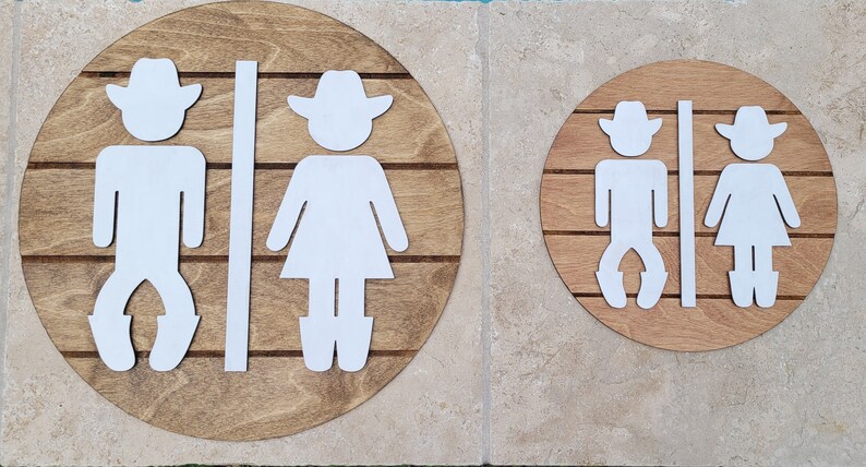May include: Two circular wooden signs featuring white cutout figures of a cowboy and a cowgirl. Each sign has horizontal grooves across the wood surface, with a vertical white divider strip separating the two figures on each circular plaque.