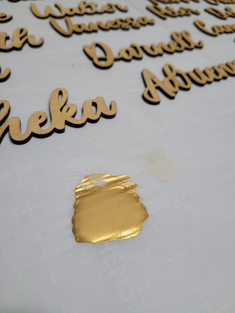 May include: Multiple wooden name cutouts in a cursive script font, featuring names such as Walter, Vanessa, Darnell, and Sheka, arranged on a white surface with a single, distinct smear of metallic gold paint positioned in the foreground.