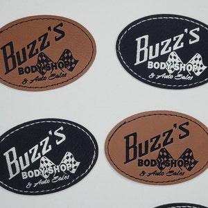 May include: Four oval-shaped leather patches with black stitching. Each patch has the text "Buzz's Body Shop & Auto Sales" in black lettering with a checkered flag graphic. Two patches are brown leather with black lettering and two patches are black leather with silver lettering.