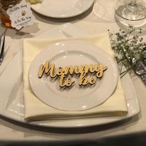 May include: A light-colored wooden cutout sign featuring the cursive text Mommy to be, placed on a white ceramic plate. The plate rests on a folded cream-colored napkin, which is centered on a larger white square plate with rounded corners.