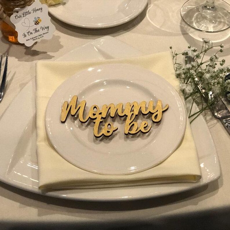 May include: A light-colored wooden cutout sign featuring the cursive text Mommy to be, placed on a white ceramic plate. The plate rests on a folded cream-colored napkin, which is centered on a larger white square plate with rounded corners.