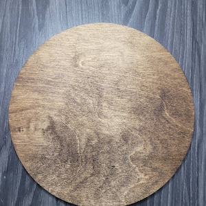 May include: A circular wooden plaque featuring a medium brown stain that highlights the natural grain patterns of the wood. The surface is smooth and flat, showcasing a consistent circular shape with visible wood texture throughout the piece.