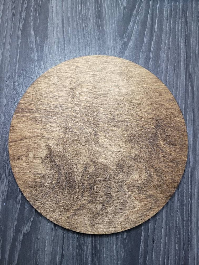 May include: A circular wooden plaque featuring a medium brown stain that highlights the natural grain patterns of the wood. The surface is smooth and flat, showcasing a consistent circular shape with visible wood texture throughout the piece.