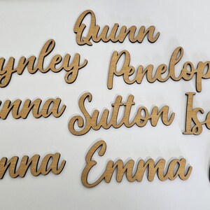 May include: A collection of laser-cut wooden name signs featuring the names Quinn, Brynley, Penelope, Lilyanna, Sutton, Isabelle, McKenna, Emma, and Greta. Each name is crafted in a cursive script font with a natural light brown wood finish.