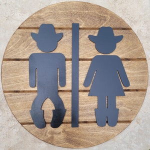May include: A circular wooden sign with a medium brown stained finish and horizontal grooves. Mounted on the surface are two black silhouettes of figures wearing cowboy hats, separated by a vertical black bar, representing restroom signage.