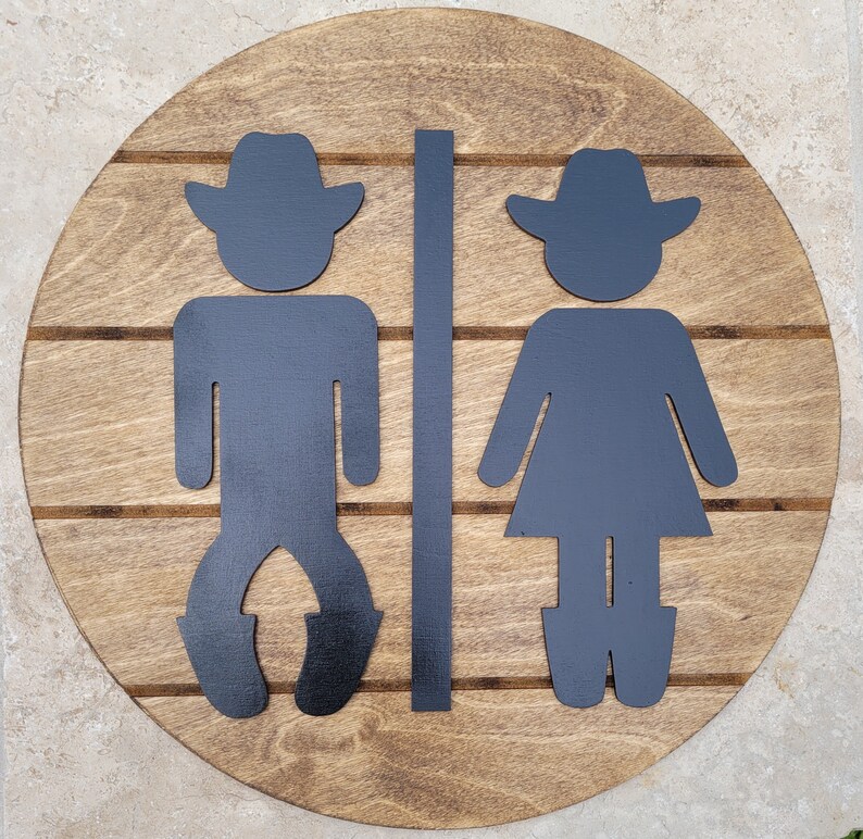 May include: A circular wooden sign with a medium brown stained finish and horizontal grooves. Mounted on the surface are two black silhouettes of figures wearing cowboy hats, separated by a vertical black bar, representing restroom signage.