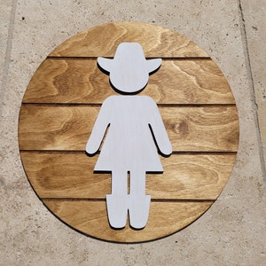 May include: A circular wooden sign featuring a dark brown stained finish with horizontal grooves. A white cutout silhouette of a figure wearing a cowboy hat and boots is centered on the wood, creating a contrast between the light and dark elements.