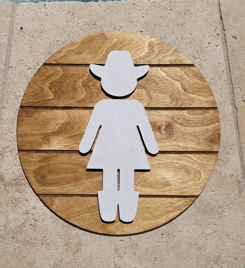 May include: A circular wooden sign featuring a dark brown stained finish with horizontal grooves. A white cutout silhouette of a figure wearing a cowboy hat and boots is centered on the wood, creating a contrast between the light and dark elements.