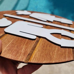 May include: A circular wooden sign featuring white, flat, laser-cut silhouettes of two figures separated by a horizontal line. The wood has a visible grain with a medium-brown stain, and the figures are arranged in a stylized, minimalist graphic design.