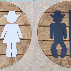 May include: Two circular wooden signs featuring horizontal slats and cutout figures. The left sign displays white cowboy and cowgirl silhouettes separated by a vertical bar. The right sign features identical black silhouettes on a matching wood base.