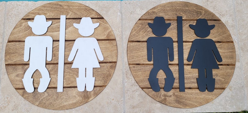 May include: Two circular wooden signs featuring horizontal slats and cutout figures. The left sign displays white cowboy and cowgirl silhouettes separated by a vertical bar. The right sign features identical black silhouettes on a matching wood base.