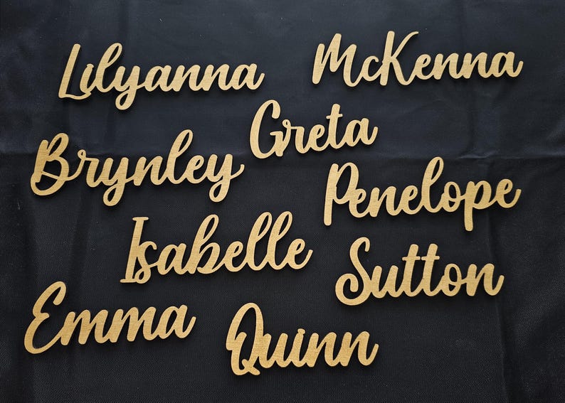 May include: A collection of gold-colored, script-style cutouts of the names Lilyanna, McKenna, Brynley, Greta, Penelope, Isabelle, Sutton, Emma, and Quinn. Each name is crafted in a cursive font and features a smooth, metallic gold finish.