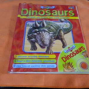 May include: A red book with a plastic cover featuring a green dinosaur on the front. The book title is "Dinosaurs" and the subtitle is "The book and CD-ROM that work together". The book cover also features a CD with the text "Dinosaurs" on it.