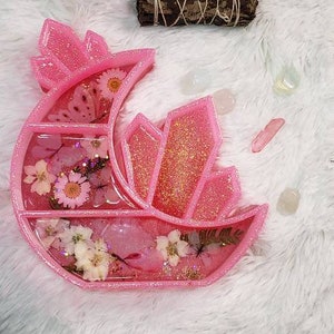 May include: Pink resin moon-shaped trinket dish with glitter and dried flowers. The dish has multiple compartments and is perfect for holding small items like jewelry or crystals.