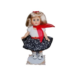 Pu&ograve; includere: Una bambola vintage con capelli biondi, indossa un top bianco, una gonna a fantasia bianca e nera e una sciarpa rossa. La bambola ha gli occhi blu e si trova su una base decorativa, mostrando un'estetica retr&ograve;.