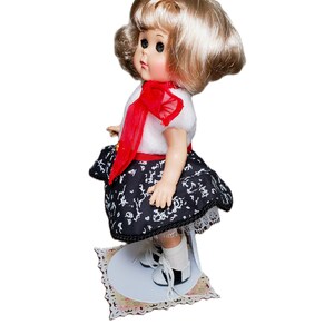 Pu&ograve; includere: Una bambola vintage con capelli biondi, indossa un top bianco con un fiocco rosso, una gonna a fantasia bianca e nera e scarpe bianche e nere. La bambola &egrave; in piedi su un supporto decorativo con un motivo floreale.