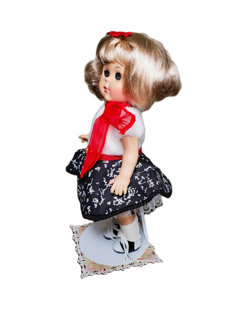 Pu&ograve; includere: Una bambola vintage con capelli biondi, indossa un top bianco con un fiocco rosso, una gonna a fantasia bianca e nera e scarpe bianche e nere. La bambola &egrave; in piedi su un supporto decorativo con un motivo floreale.
