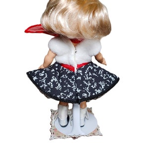 Pu&ograve; includere: Una bambola vintage, vista da dietro, con capelli biondi e sciarpa rossa. La bambola indossa un top bianco, una gonna fantasia bianca e nera e scarpe bianche. Montata su un supporto bianco con base decorativa.