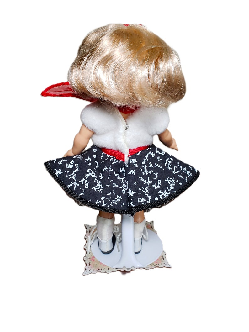 Pu&ograve; includere: Una bambola vintage, vista da dietro, con capelli biondi e sciarpa rossa. La bambola indossa un top bianco, una gonna fantasia bianca e nera e scarpe bianche. Montata su un supporto bianco con base decorativa.