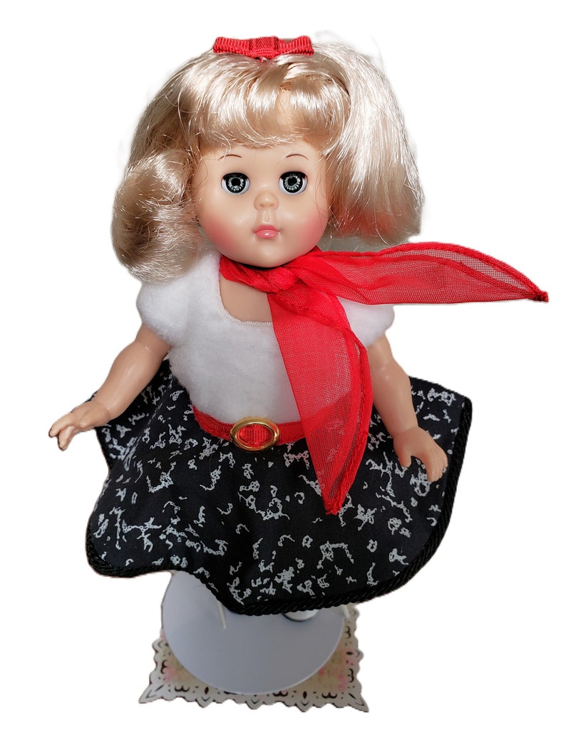 Pu&ograve; includere: Bambola vintage con capelli biondi, indossa un top bianco, una gonna nera con un motivo bianco e una sciarpa rossa. La bambola ha un fiocco rosso e una cintura rossa con fibbia dorata. La bambola &egrave; su un supporto bianco.