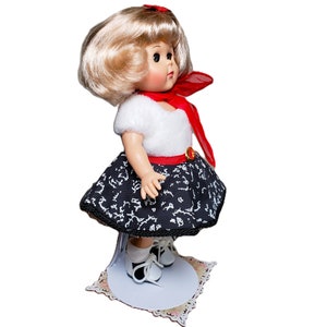Pu&ograve; includere: Una bambola vintage con capelli biondi, indossa un top bianco, una gonna a fantasia bianca e nera e scarpe bianche. La bambola ha una sciarpa rossa e una cintura rossa. &Egrave; esposta su un supporto bianco con una base decorativa.