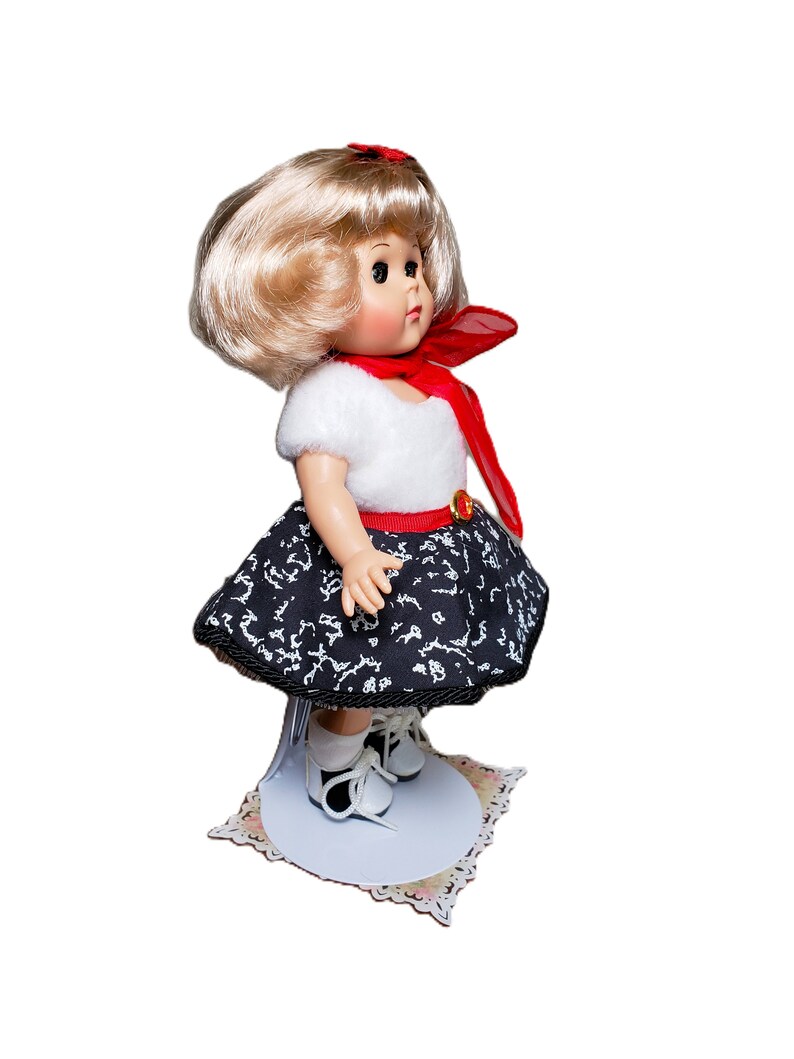 Pu&ograve; includere: Una bambola vintage con capelli biondi, indossa un top bianco, una gonna a fantasia bianca e nera e scarpe bianche. La bambola ha una sciarpa rossa e una cintura rossa. &Egrave; esposta su un supporto bianco con una base decorativa.