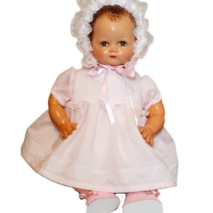 May include: A vintage-style doll with brown hair, blue eyes, and red lips, wearing a pale pink dress with short sleeves and white lace trim. The doll is adorned with a matching white lace bonnet tied with a pink ribbon and pink shoes with bows.
