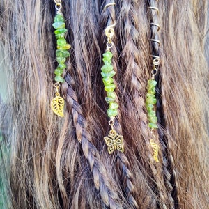 May include: Three gold hair beads with green gemstone beads and gold charms. The charms are a leaf, a butterfly, and a small leaf. The beads are strung on wire and are attached to the hair with a small loop.