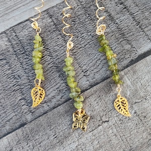May include: Three gold-colored hair spirals with green gemstone beads and gold charms. One charm is a butterfly, and two are leaf-shaped. The hair accessories are displayed on a gray wooden surface.