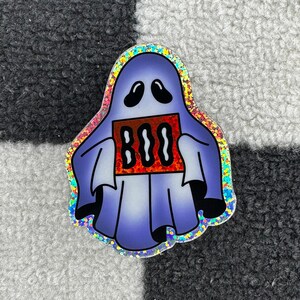 May include: A holographic sticker of a purple ghost holding a red sign that says "BOO" in a wavy font.