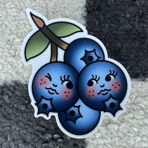 May include: A sticker of three blue blueberries with cute faces. The blueberries are arranged in a cluster with a green stem and leaves.