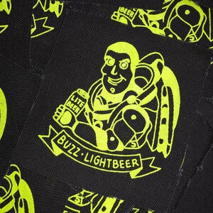 May include: A black and yellow patch with a cartoon image of Buzz Lightyear holding a can of "Lite Beer" and a banner that reads "Buzz Lightbeer".