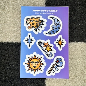 May include: A sticker sheet with a purple background featuring various illustrations, including a yellow and orange tiger head, a blue crescent moon, a yellow sun with a face, a blue safety pin with two stars, and a match with blue smoke. The text "MOON DUST GIRLS" and "filler sticker sheet #2" is printed at the top of the sheet.