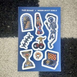 May include: A sheet of stickers featuring various illustrations, including a skeleton dancing, a lava lamp, a cherry with two cherries, a barbed wire, a tooth, an hourglass, and a record player. The stickers are on a blue background with the text "Tufti Broad + Moon Dust Girls".