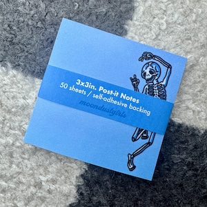 May include: A light blue square of Post-it Notes with a blue band that reads "3x3in. Post-it Notes, 50 sheets / self-adhesive backing, moondustgirls." The notes have a black and white skeleton illustration.