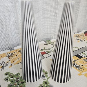 Peut inclure: Deux vases coniques et hauts avec un motif à rayures noires et blanches. Les vases sont présentés sur une nappe à motifs avec un rideau blanc en arrière-plan. Les vases mesurent environ 25 cm de haut.