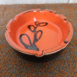 May include: Orange ceramic ashtray with a black floral design. The ashtray has a scalloped edge and a small lip for resting cigarettes. The interior is a glossy orange, and the design is in the center.