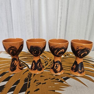 May include: Four ceramic goblets with a warm orange glaze and black abstract designs. The goblets have a flared top and a pedestal base. They are arranged on a patterned tablecloth with a neutral background.