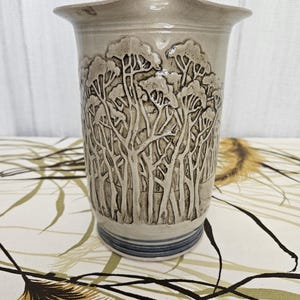 May include: A beige ceramic vase with a forest design carved into the exterior. The vase has a flared top with a unique shape and a blue stripe near the base. The forest scene features detailed tree trunks and foliage.