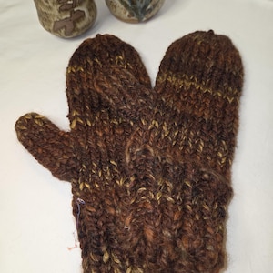 May include: A pair of brown and yellow knitted mittens. The mittens are made of thick yarn and have a textured appearance. The mittens are lying on a white surface, with two ceramic mugs in the background.